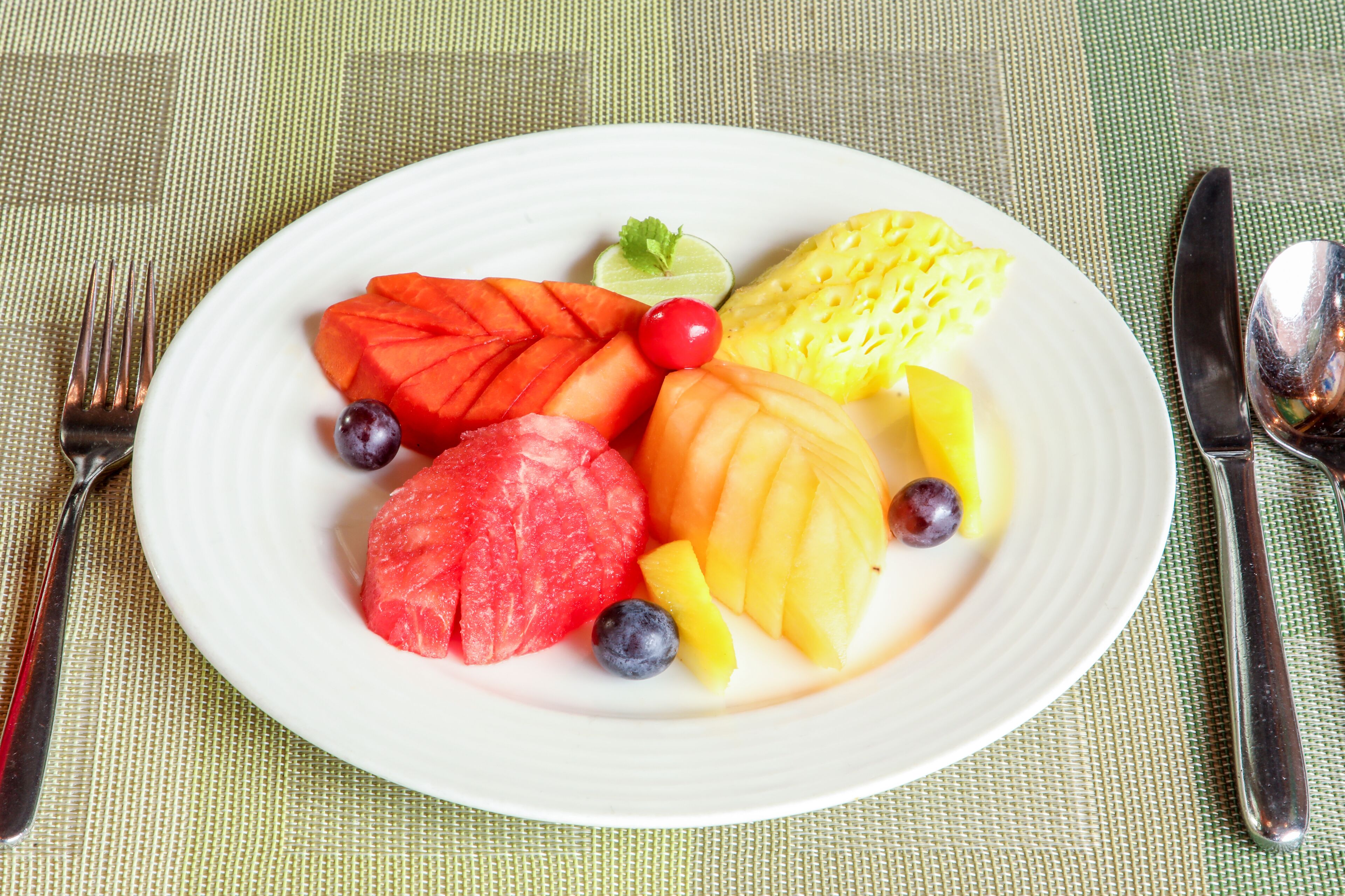 Ein Teller mit Früchten, darunter Wassermelone, Mango, Ananas und Blaubeeren, sowie ein Limettenschnitz und eine Kirsche auf weißem Geschirr.