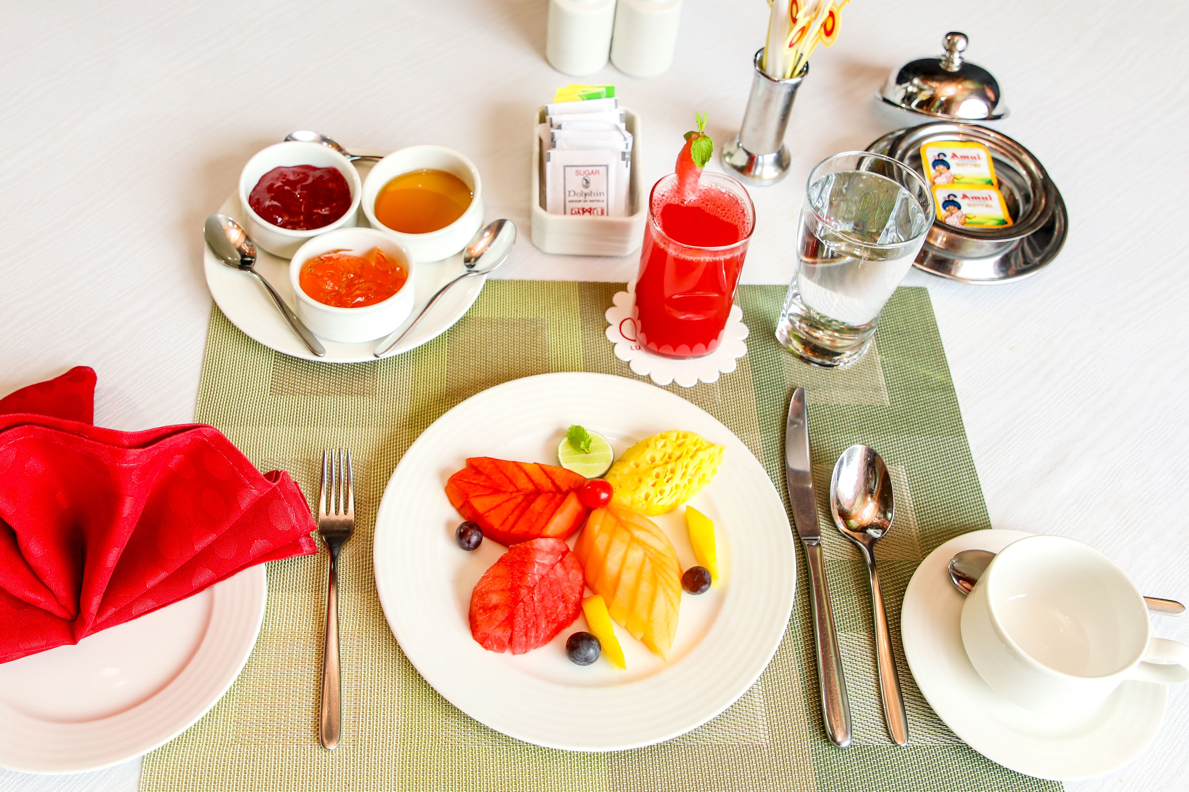A breakfast setting with a plate of fruit, a glass of red drink, and a cup of water on a table with a placemat.