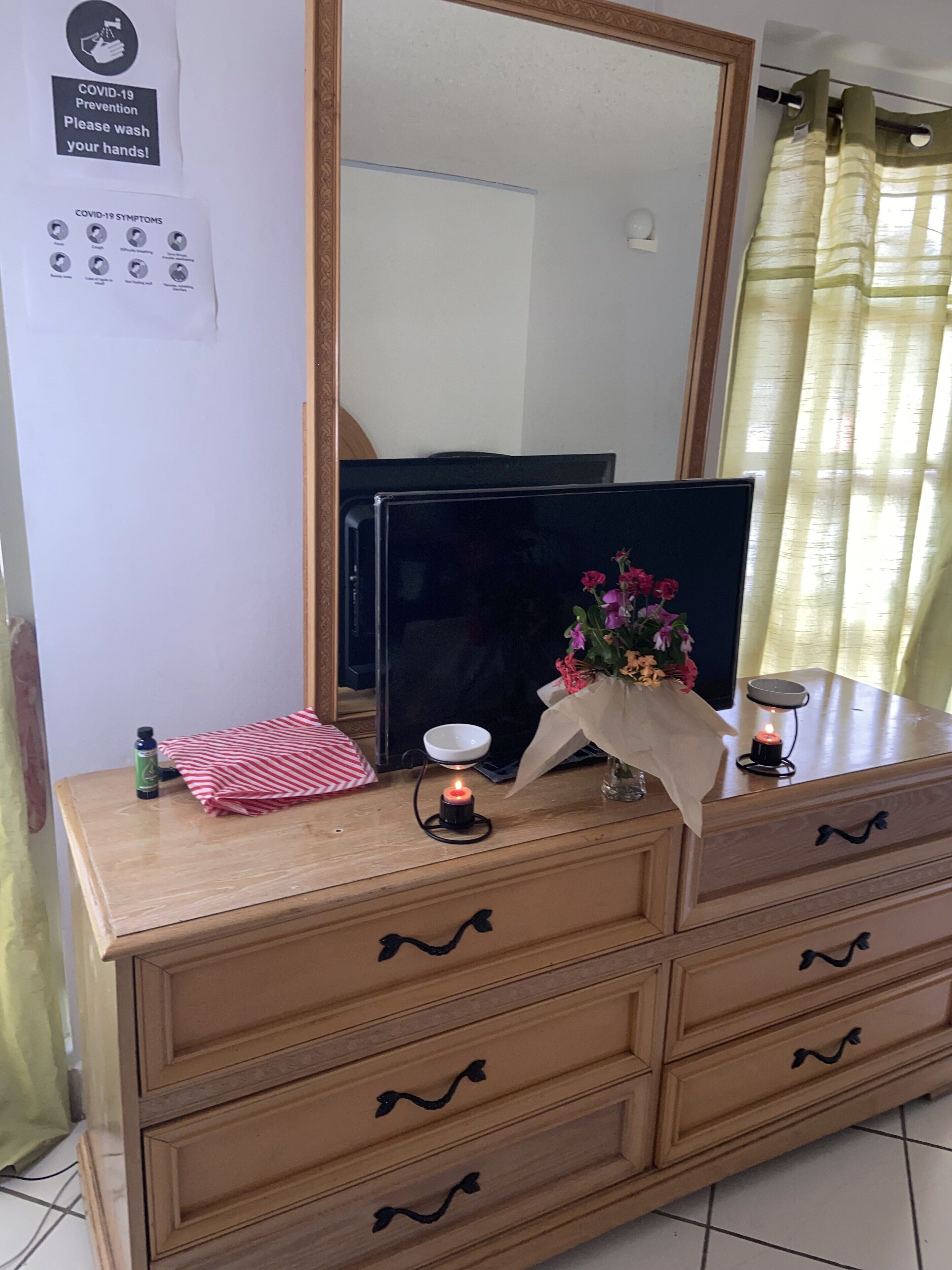 A wooden dresser with drawers, a mirror, a television, a red and white striped towel, a small bowl, a candle, and a vase with flowers.