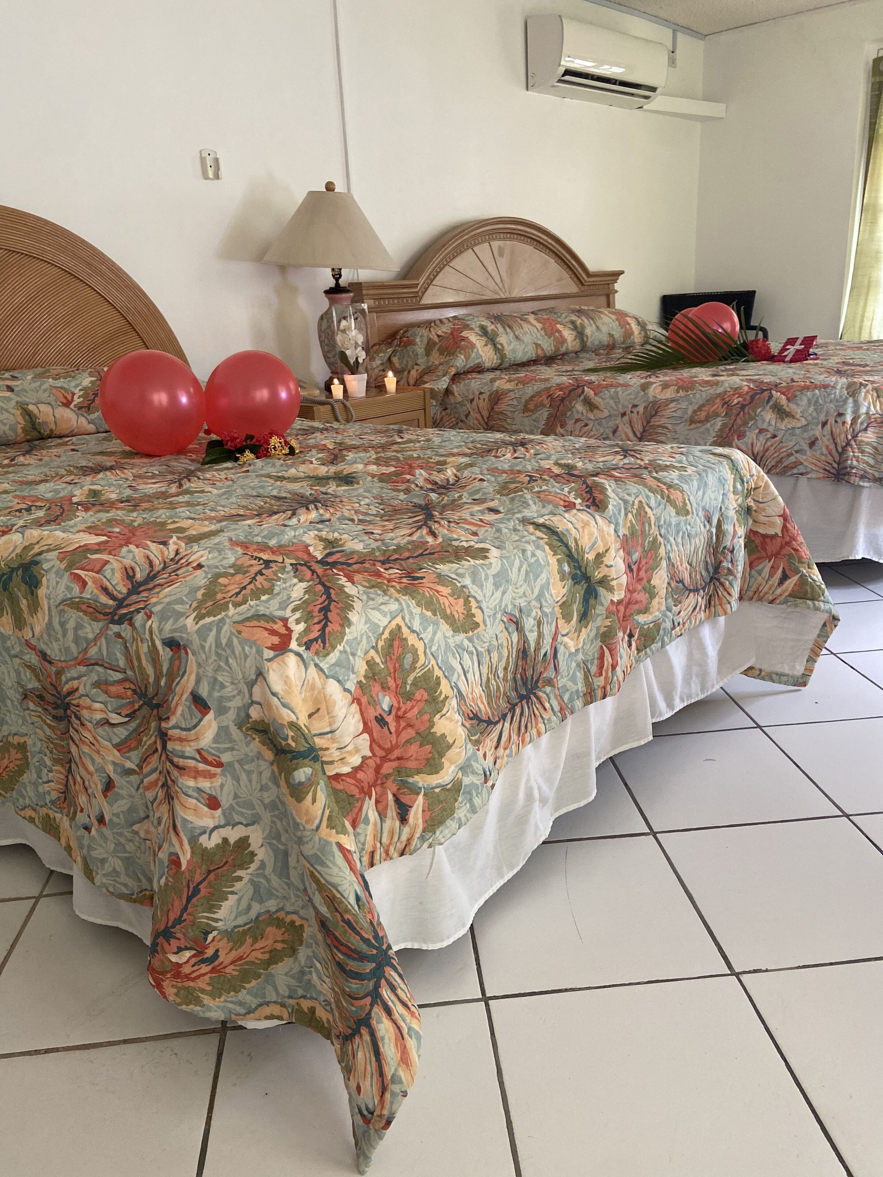 A hotel room with two beds, a wooden headboard, a wall-mounted air conditioner, and a bedside table with a lamp and decorative items.