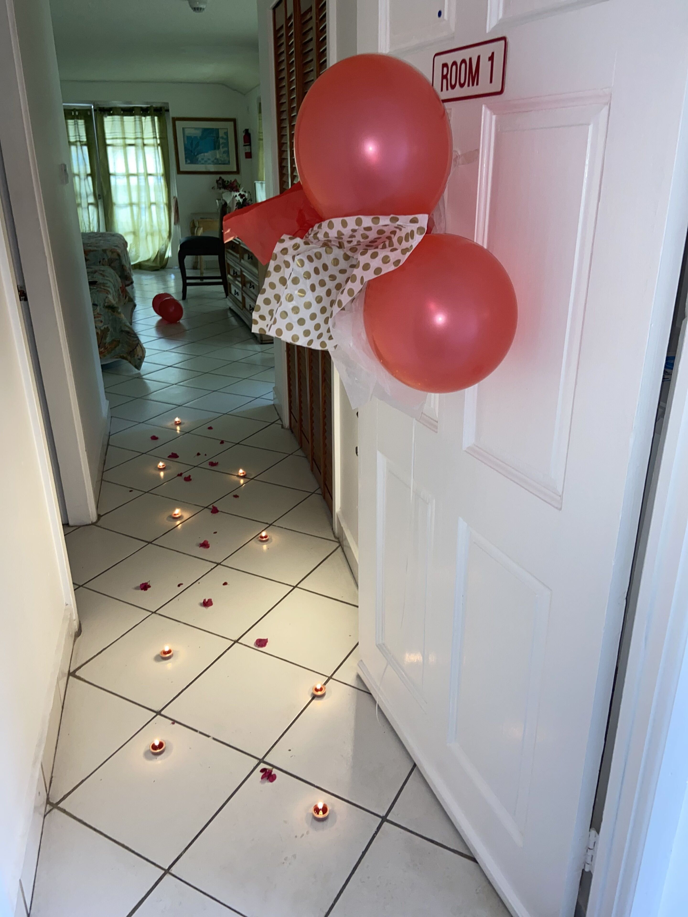 A hallway with a door labeled "ROOM 1" adorned with red balloons and a polka-dotted bow. The floor is tiled, and there are small candles and rose petals scattered around.