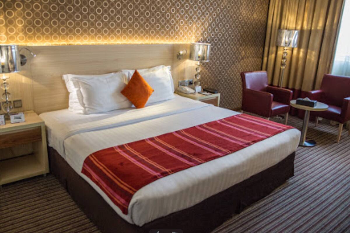 A hotel room with a large bed, a red and white striped blanket, two bedside lamps, a nightstand, a red armchair, and a small table.