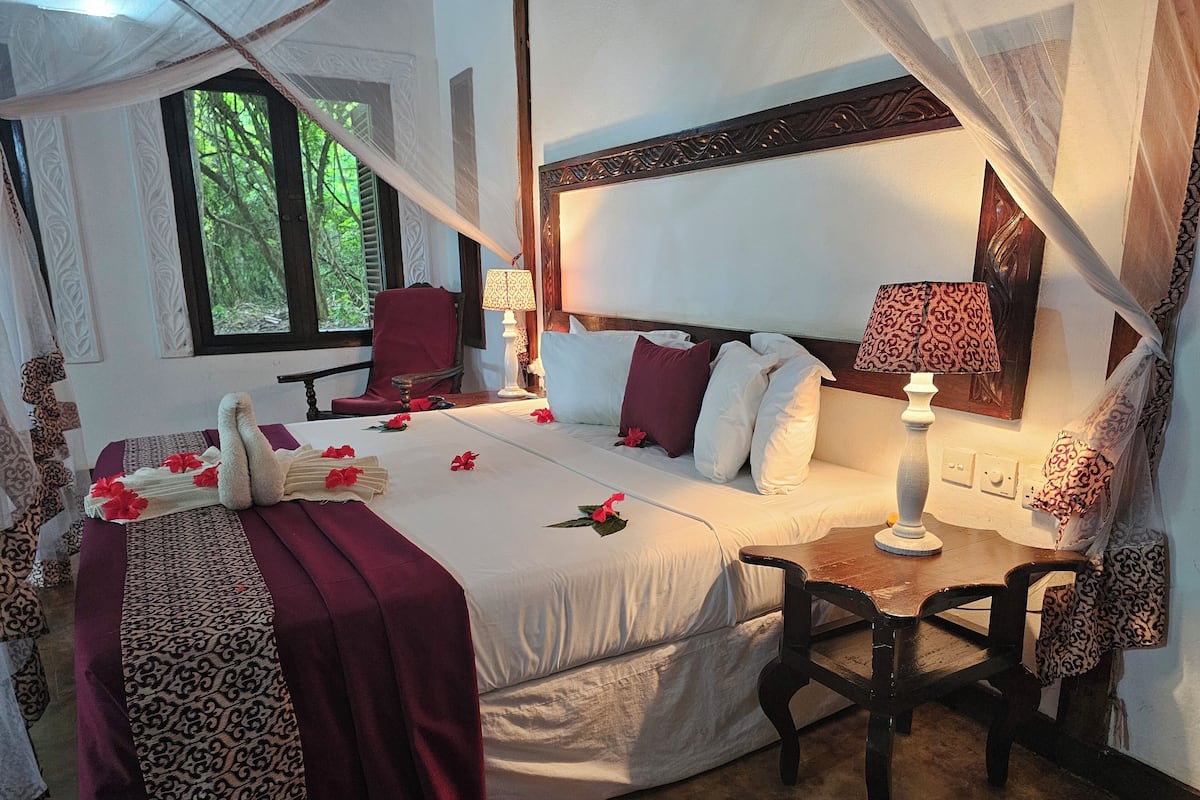 A spacious bedroom with a large bed, a sitting area with a chair, and a window with a view of greenery.