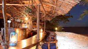 A beachside bar with wooden tables and chairs, a thatched roof, and a lantern. - Fumba Beach Lodge (Fumba)