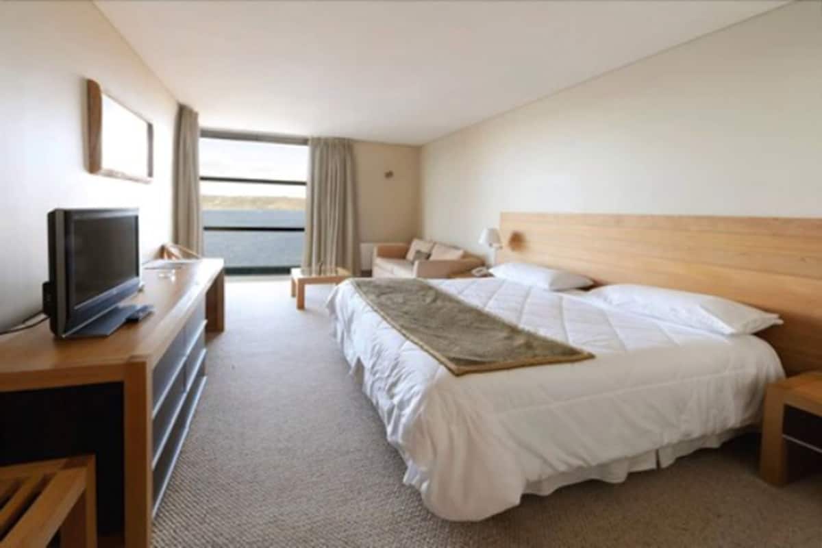 A hotel room with a bed, a desk with a television, a nightstand, and a window with curtains.