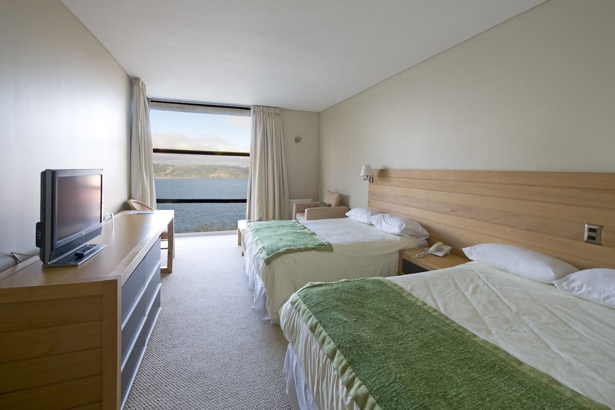 A hotel room with two beds, a TV, a desk, and a view of the sea.
