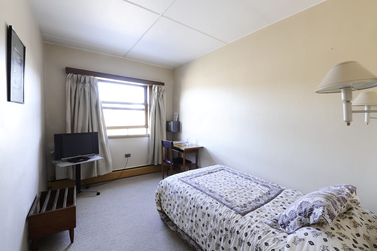 A bedroom with a bed, a desk with a computer, a TV on a stand, and a window with curtains.