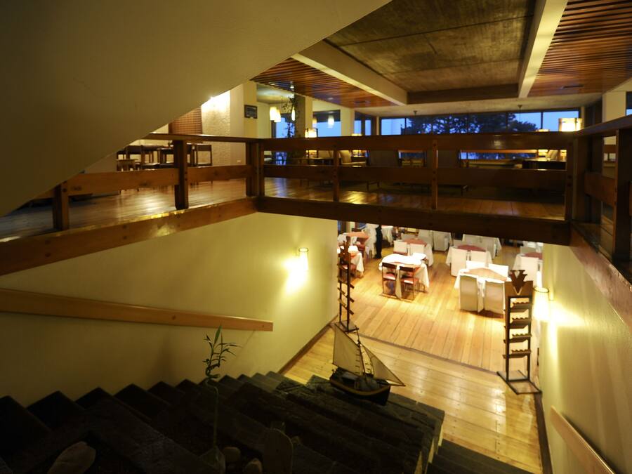 Un interior espacioso con una escalera de madera que lleva a un nivel inferior, un comedor con mesas y sillas, y vistas al exterior a través de amplios ventanales.