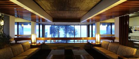 A modern lounge area with a wooden ceiling, large windows offering a view of trees, and a seating arrangement including sofas and tables.