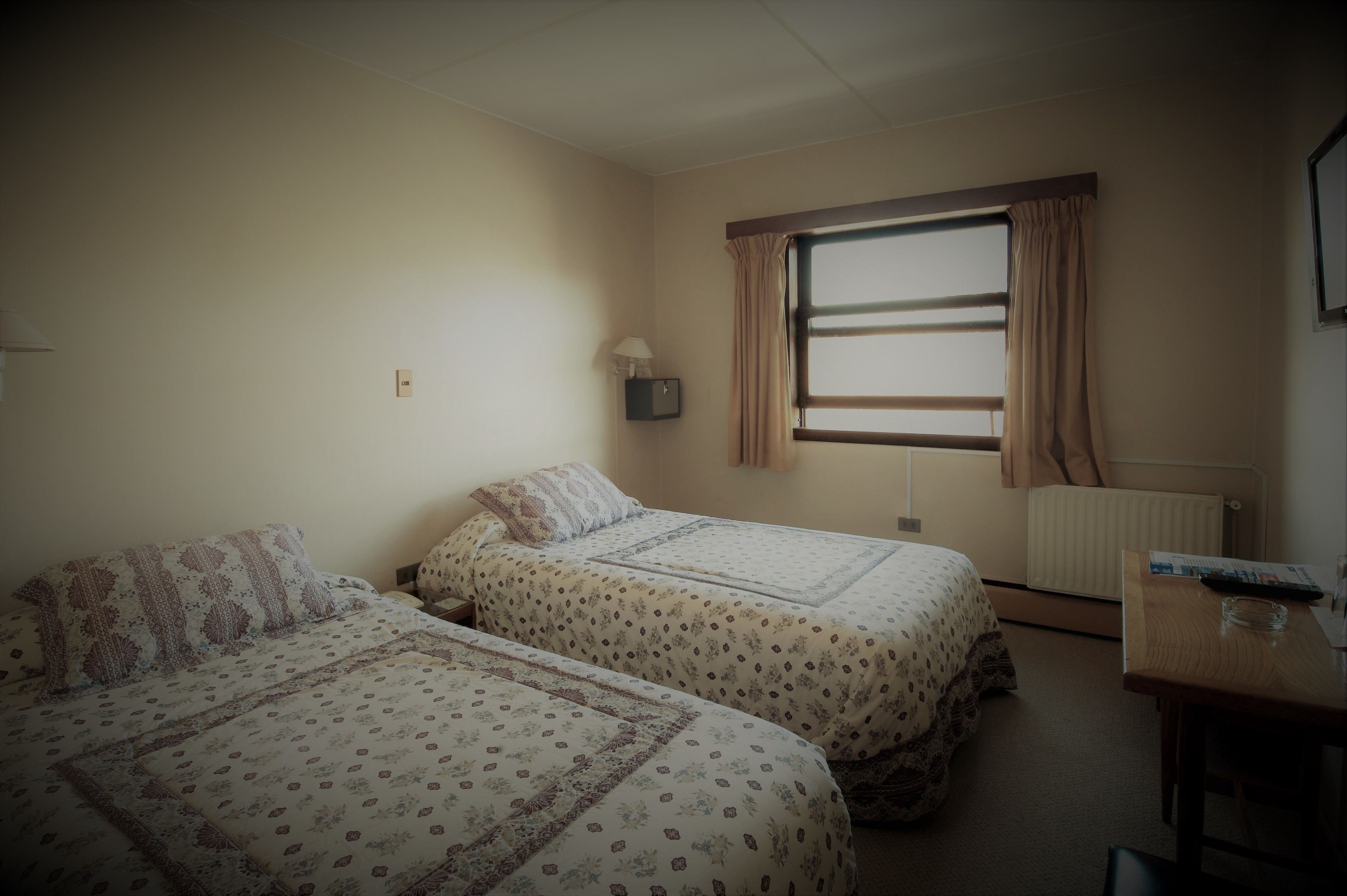 Una habitación con dos camas, una ventana con cortinas, una mesita de noche y un radiador.