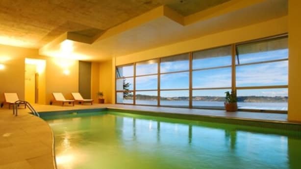 An indoor swimming pool with clear windows, lounge chairs, and a view of the water. - Hotel de Castro (Castro)