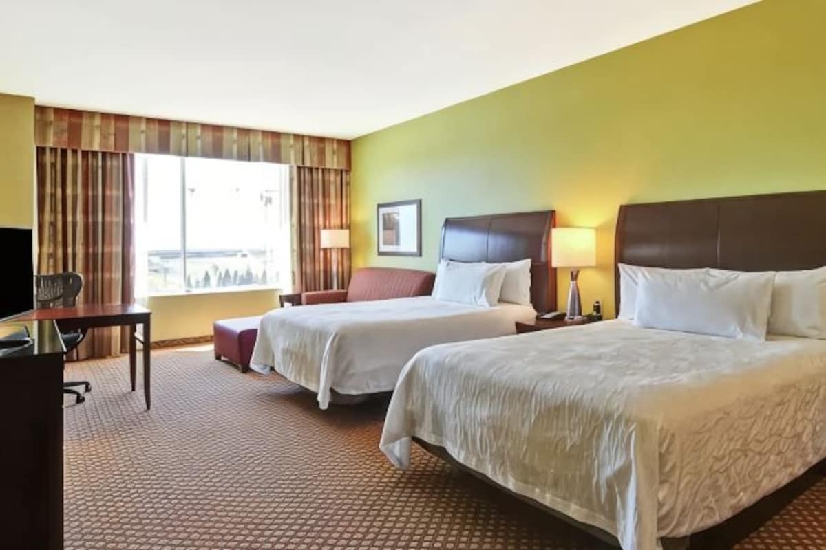 A hotel room with two beds, a desk, a chair, a TV, and a window with curtains.