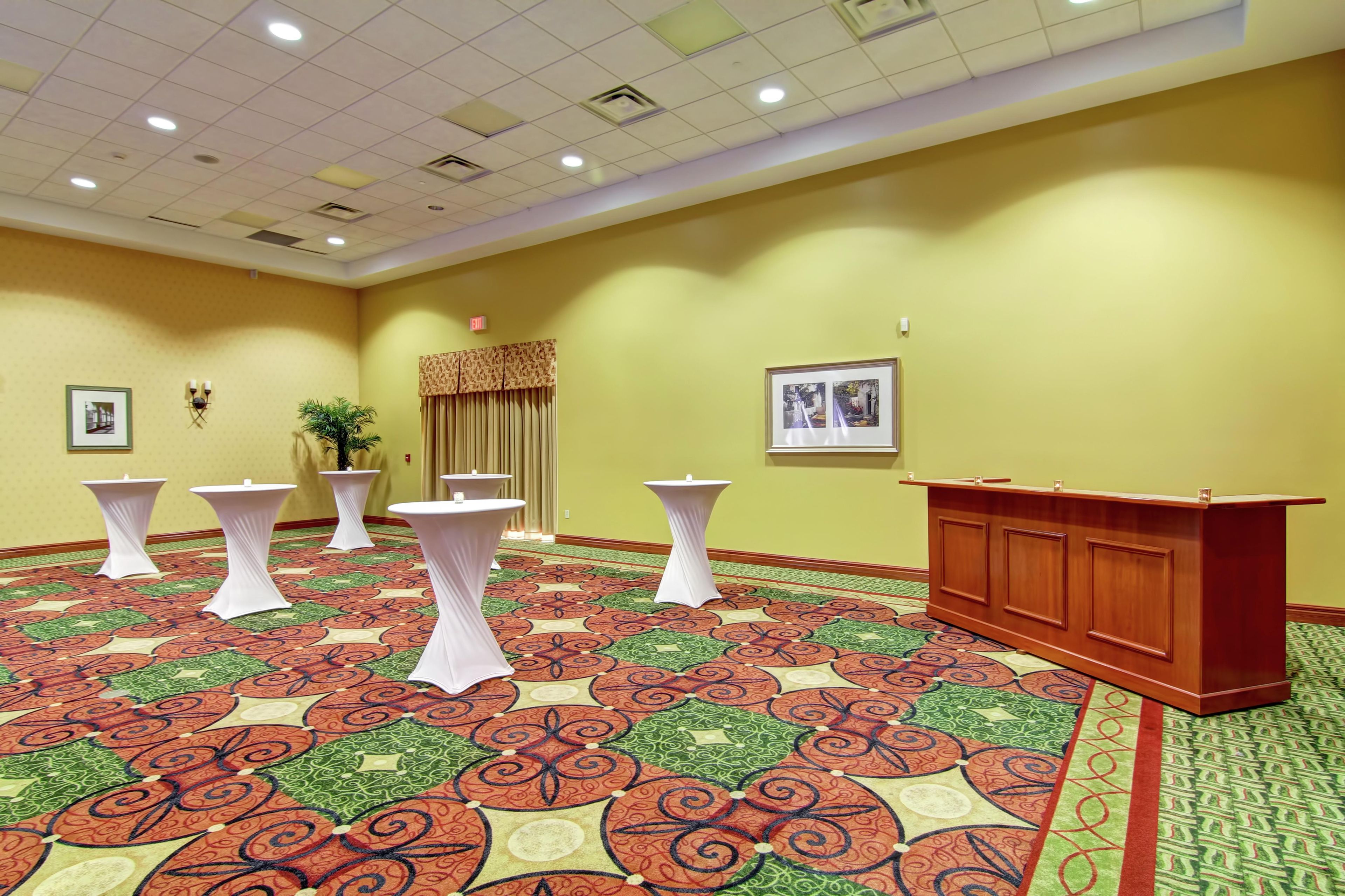 Un salón de conferencias con alfombra estampada, mesas redondas blancas, un atril de madera y una planta.