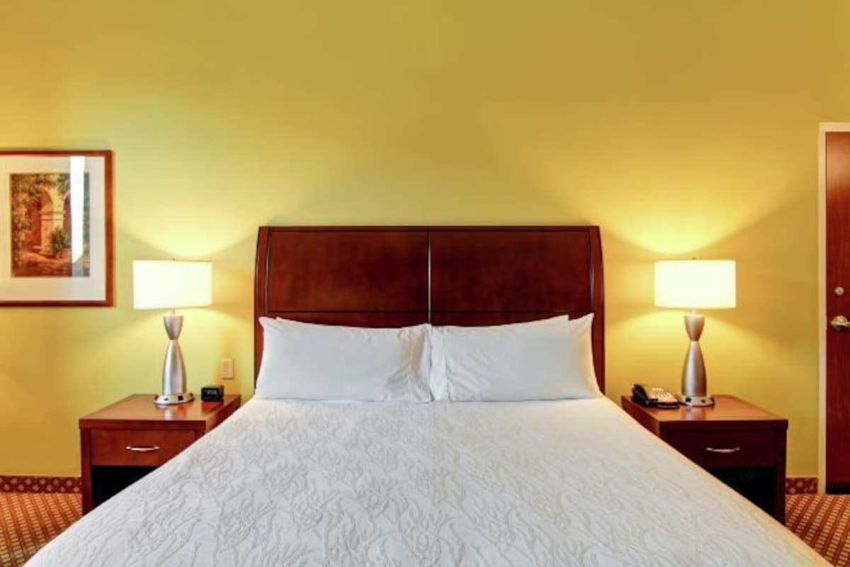A hotel room with a large bed, two bedside tables with lamps, a framed picture on the wall, and a door on the right.