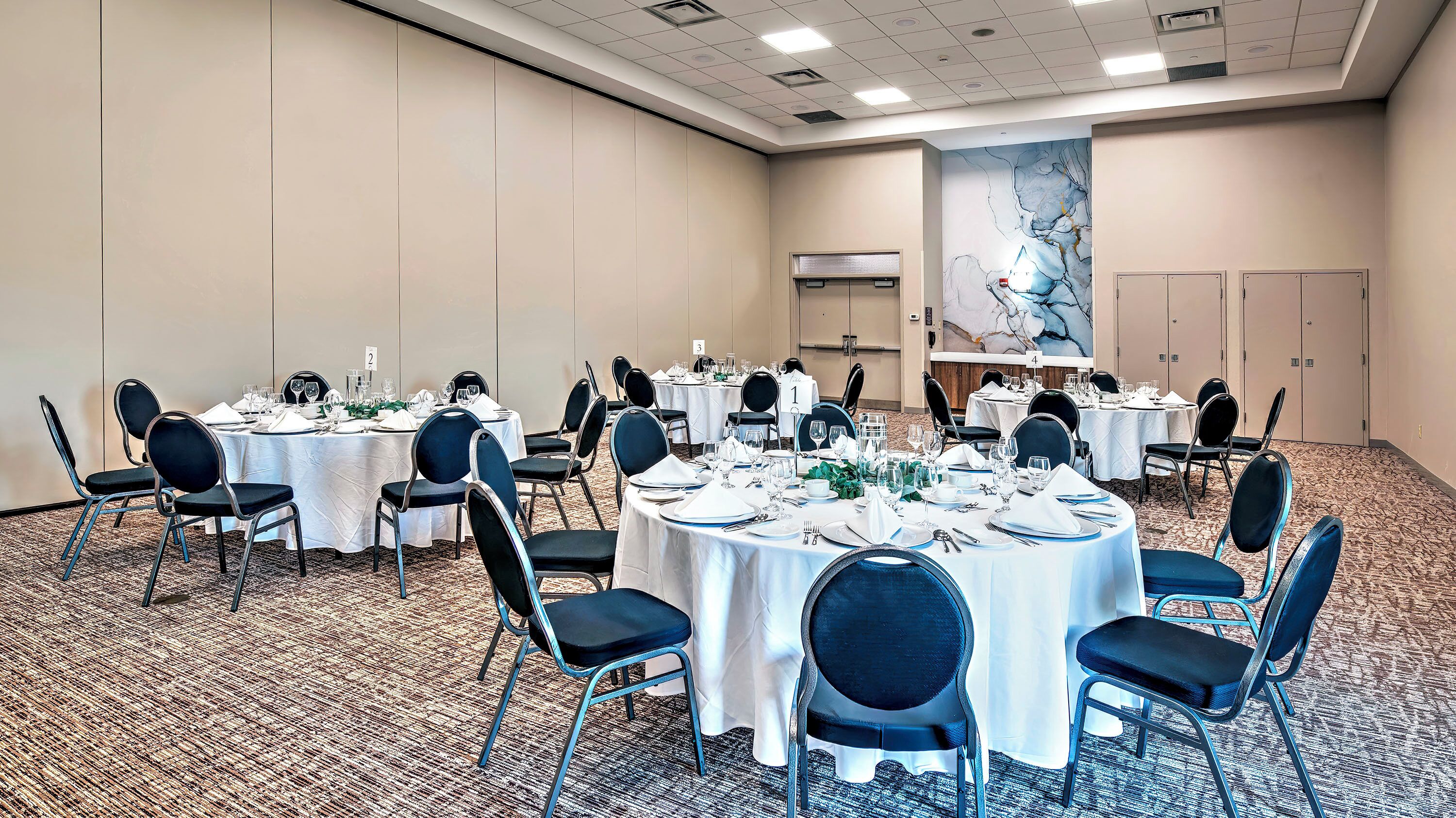 Une salle de conférence dotée de plusieurs tables rondes, chacune recouverte d’une nappe blanche et agrémentée de chaises noires et de couverts disposés avec soin.