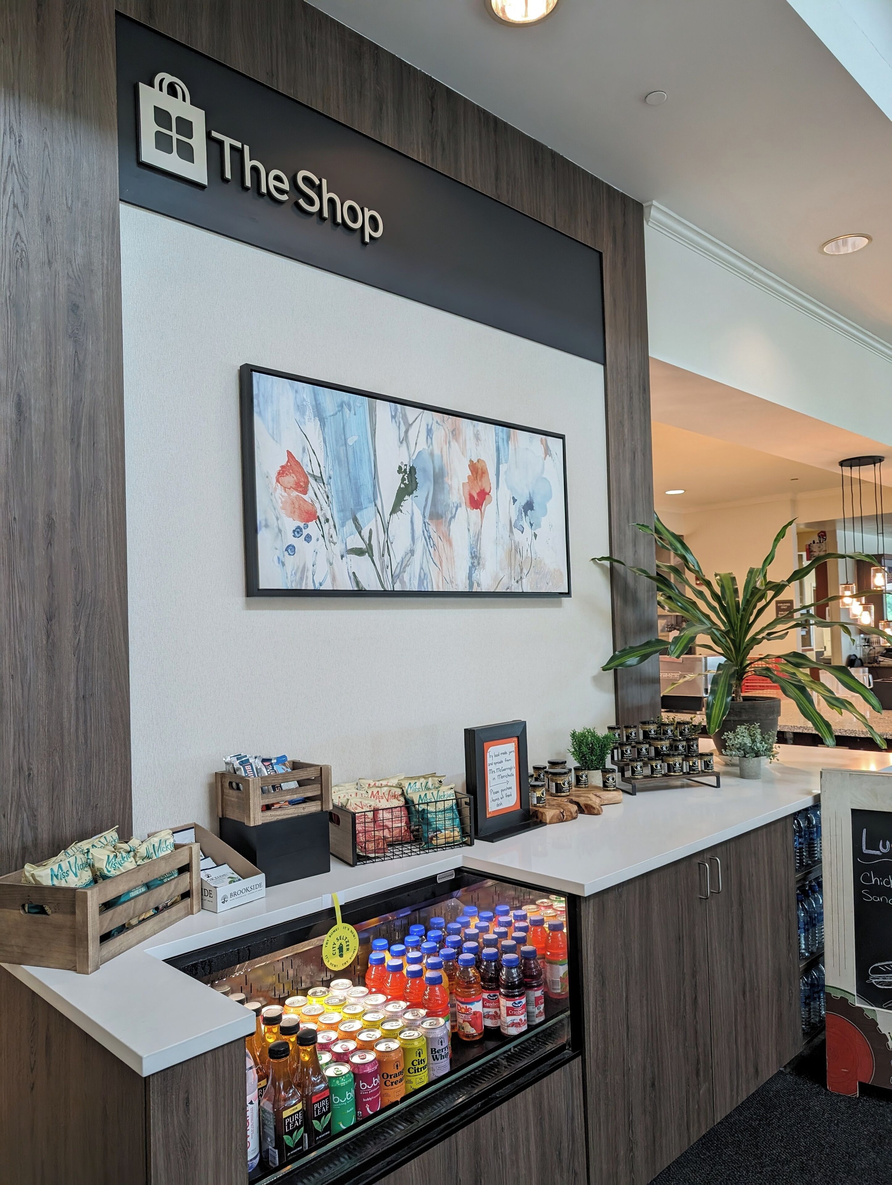 A shop counter with a sign reading "The Shop," a framed floral artwork above, and a variety of products including snacks and beverages.