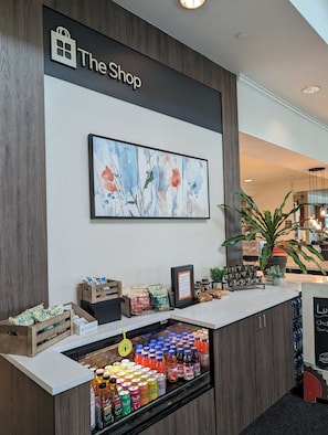 Ein Verkaufstresen mit einem Schild, auf dem "The Shop" steht, einem gerahmten Blumenbild darüber und verschiedenen Produkten, darunter Snacks und Getränke.