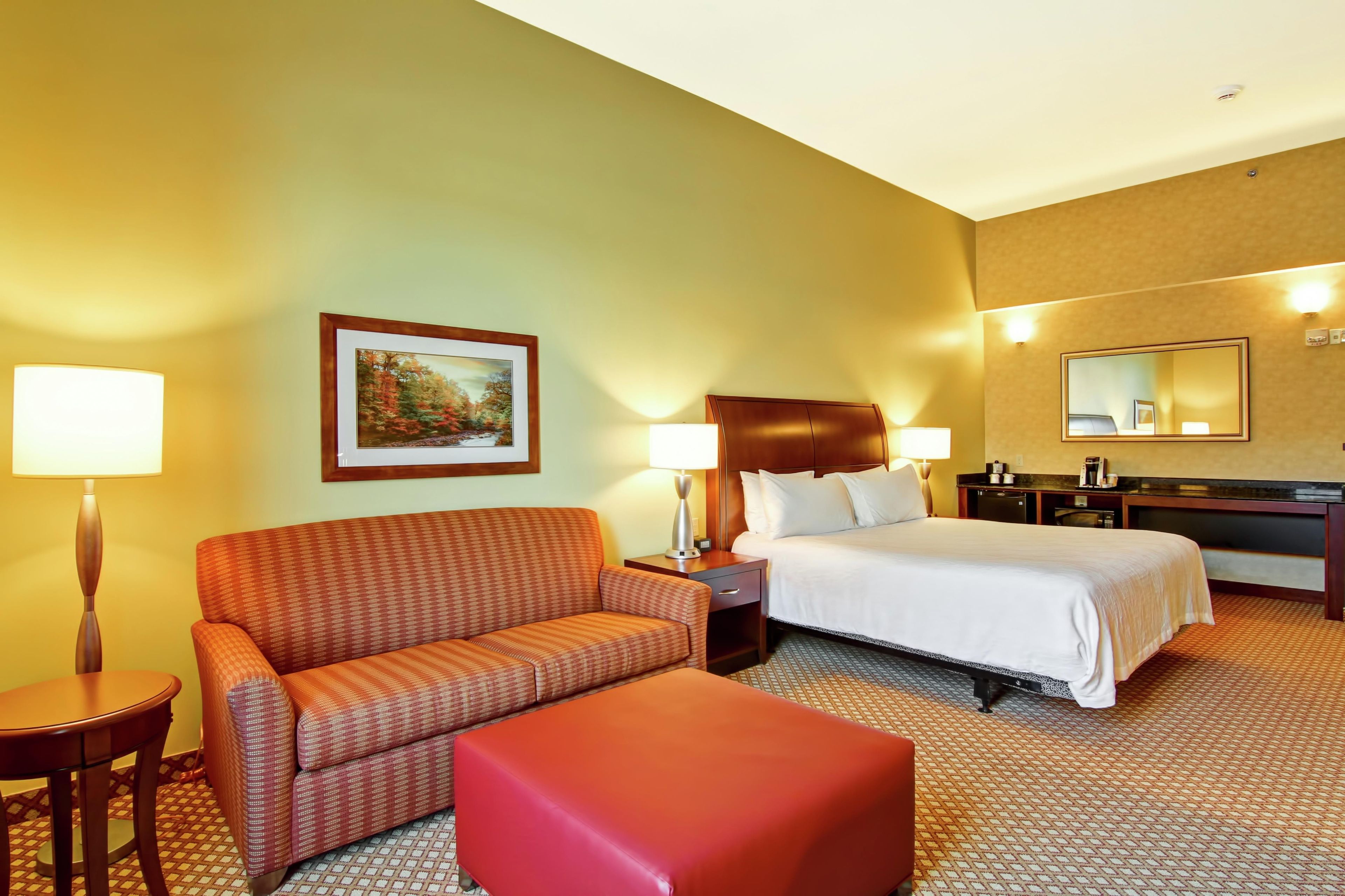 Habitación de hotel con cama, sofá, una mesita, sillón, mesita de noche, lámpara, un cuadro, espejo y escritorio con teléfono.
