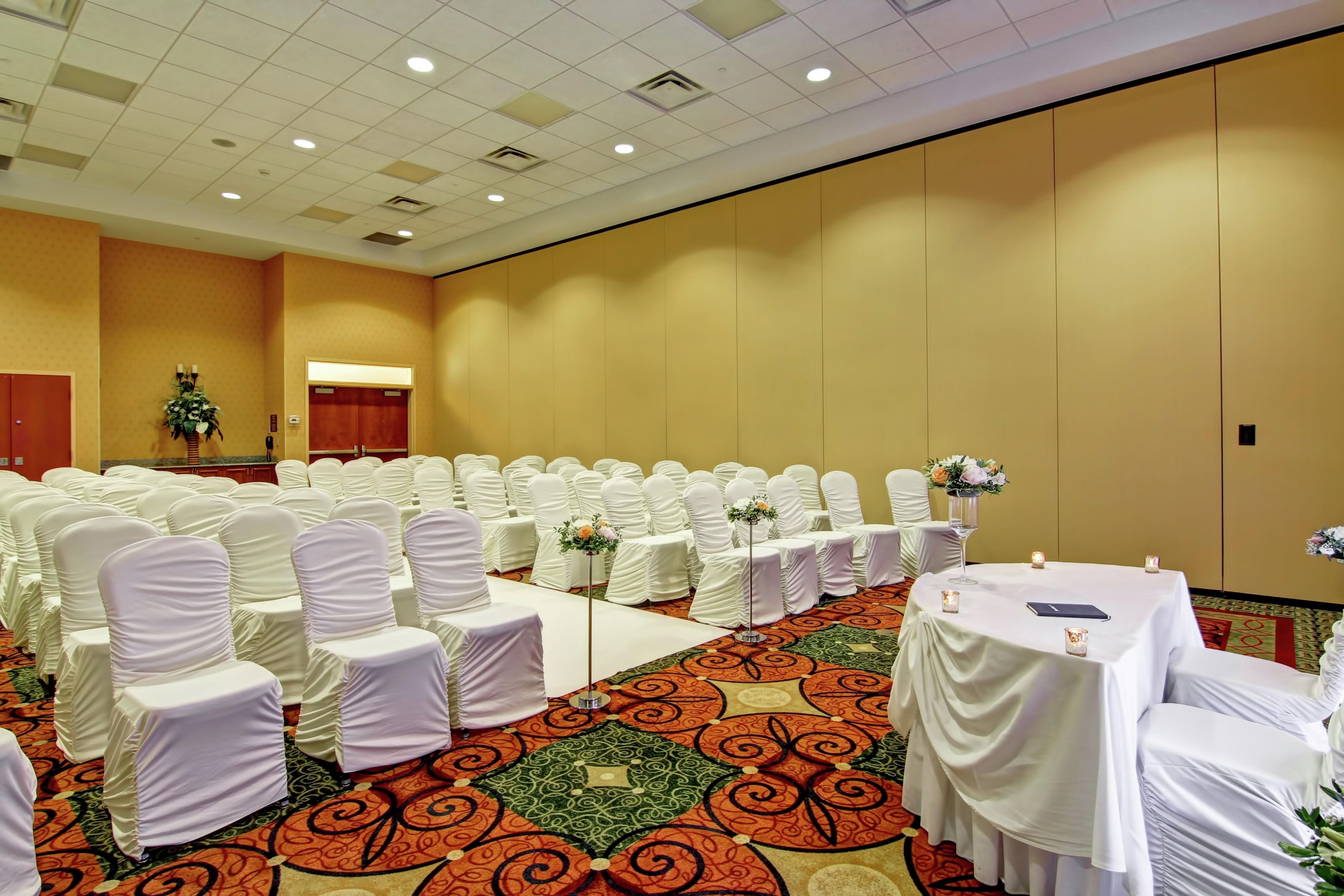 Une salle de conférence avec des rangées de chaises et de tables recouvertes de tissu blanc, une moquette à motifs et un mur sobre agrémenté d’une porte.