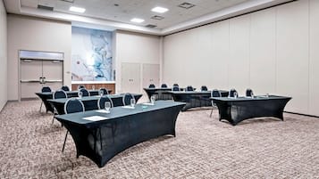 Een vergaderruimte met meerdere tafels en stoelen, een groot abstract schilderij aan de muur en een vloerbedekking.
