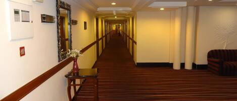 A hotel hallway with a carpeted floor, a reception desk, a mirror, and a wall-mounted phone.