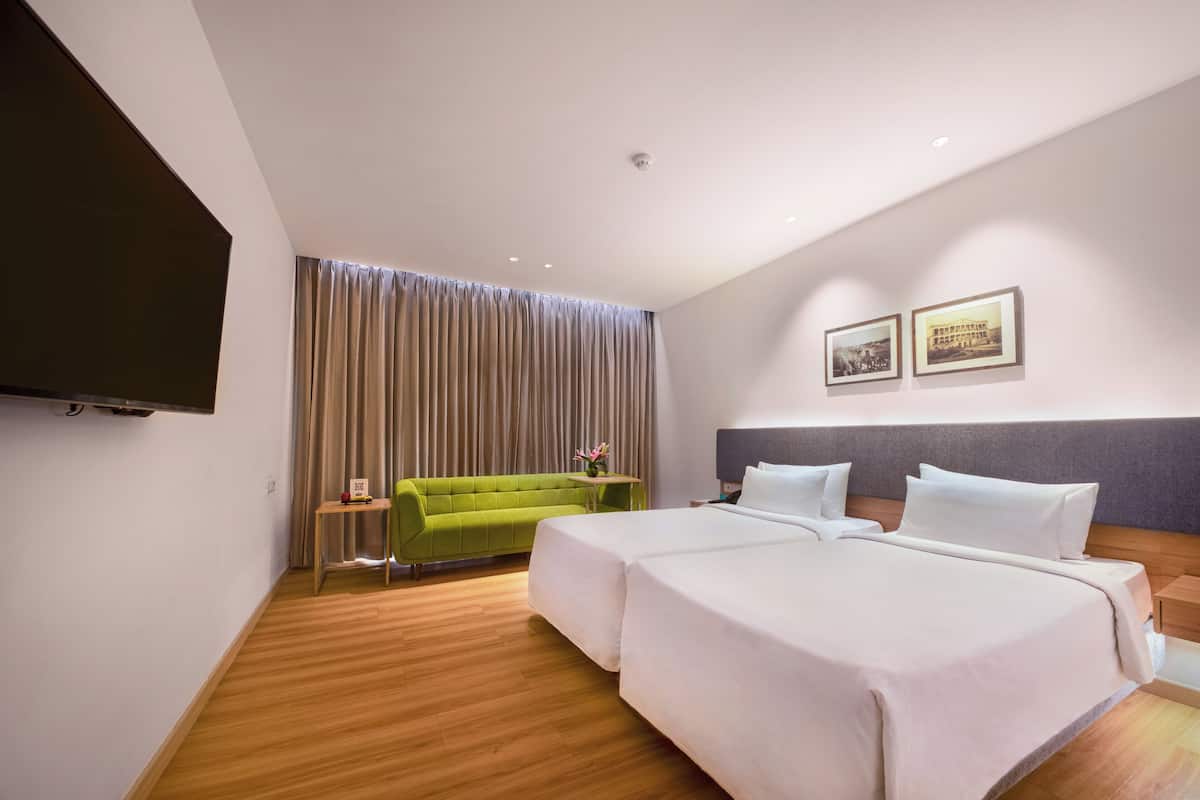 A hotel room with two beds, a green sofa, a small table with a vase, and a flat-screen TV mounted on the wall.