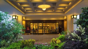 Entrada de un hotel con puerta de cristal, mostrador de recepción y un jardín ajardinado.