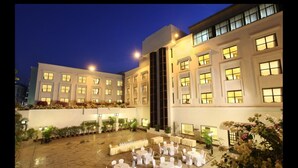 Un patio de hotel con mesas y sillas redondas, rodeado por un edificio blanco con varias ventanas y una escalera central.