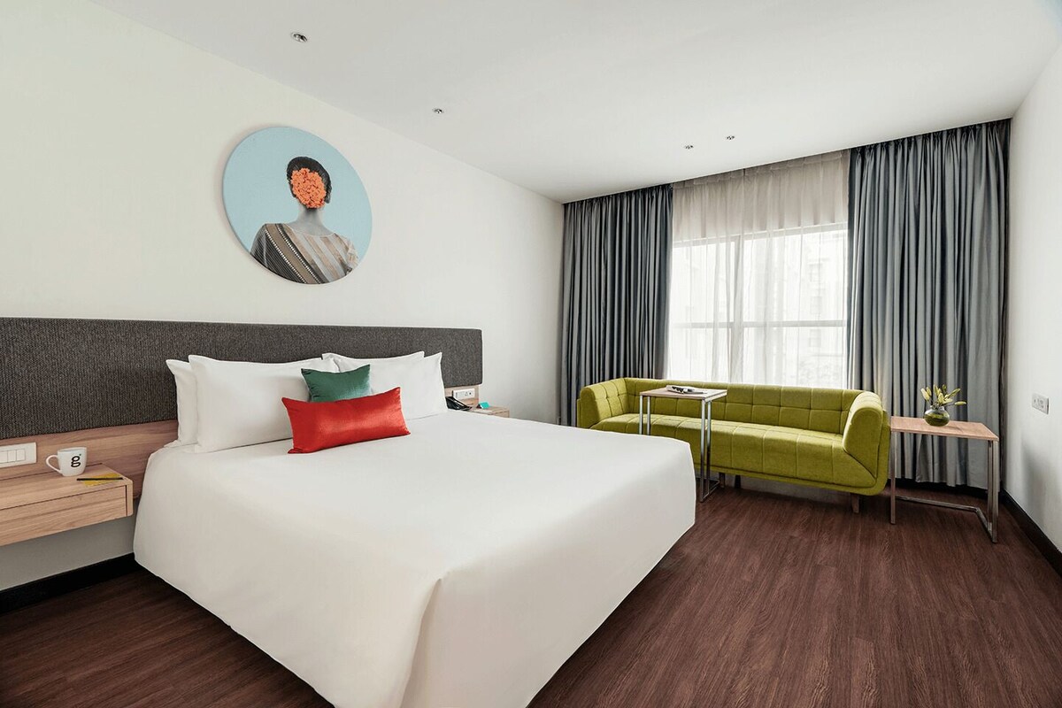 Habitación de hotel con una cama, un sofá verde, una mesita, un cuadro en la pared y una ventana con cortinas.