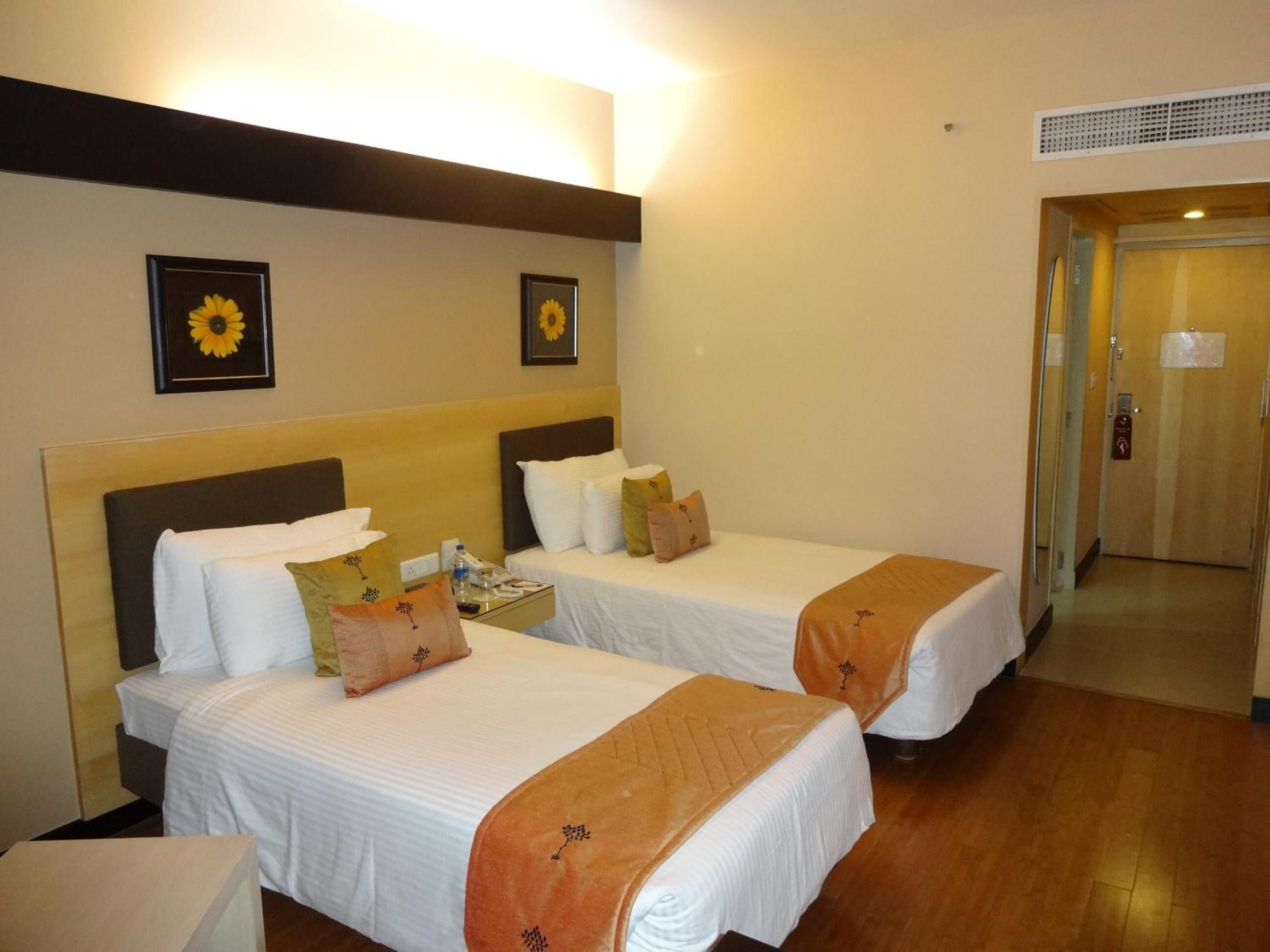 Habitación de hotel con dos camas, cada una con sábanas blancas y mantas color naranja. Hay girasoles enmarcados en la pared, una mesita de noche con un teléfono y una botella de agua, y una puerta que da a otra habitación.