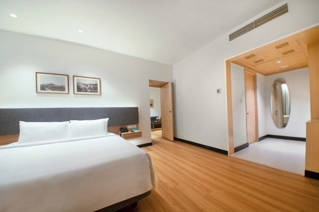 Une chambre d’hôtel moderne avec un grand lit, un plancher en bois, une tête de lit grise et une petite table de chevet.