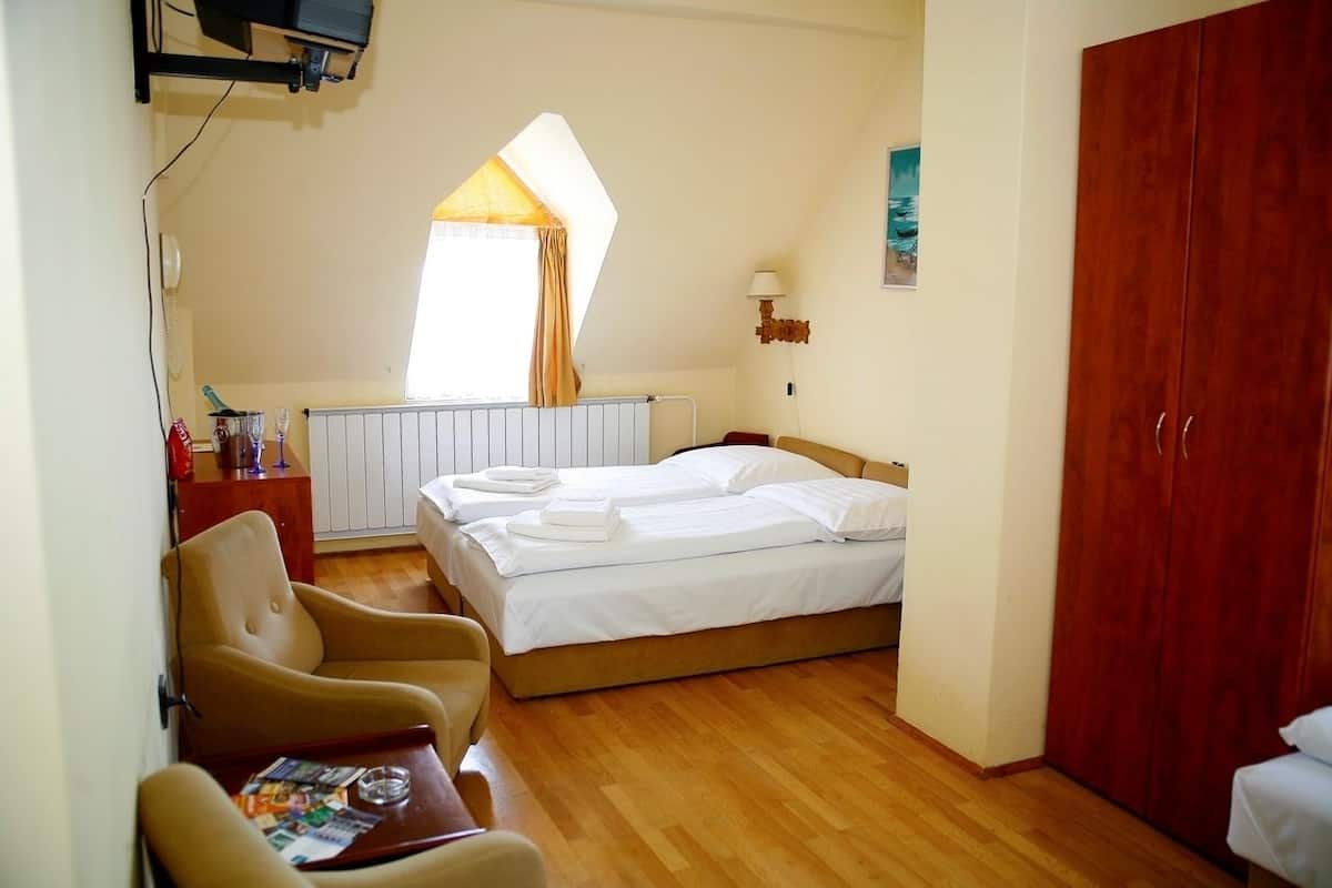 A bedroom with a bed, a chair, a table with a magazine, a radiator, a window with curtains, a lamp, and a wardrobe.