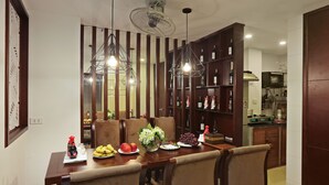 A dining area with a wooden table set for four, surrounded by chairs. There are decorative items on the table, including fruits, flowers, and a bottle. The room has wooden paneling and a ceiling fan. - Hanoi La Vision Hotel (Hanoi)