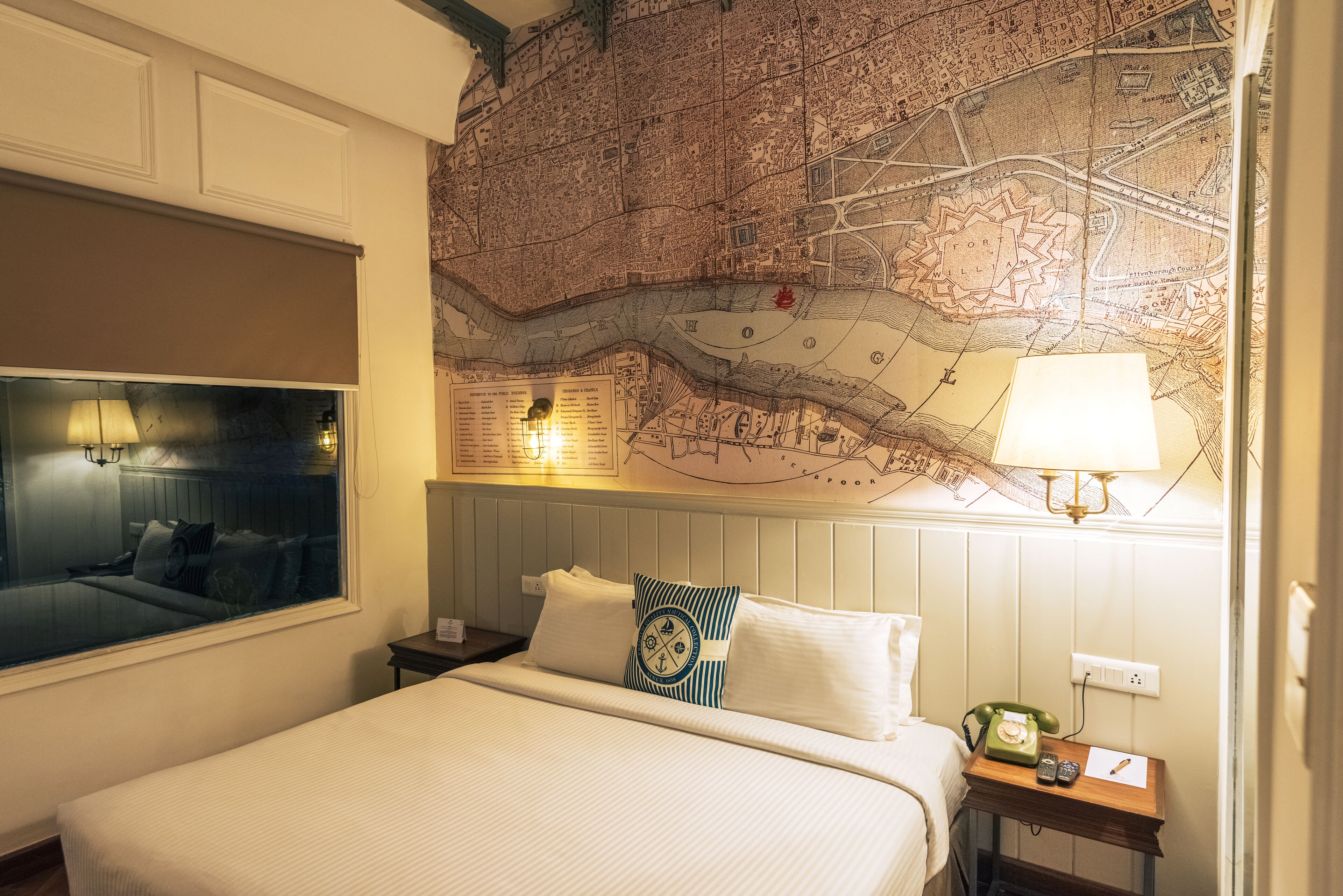 Habitación de hotel con un mapa grande en la pared, una cama con ropa blanca, una mesita de noche con una lámpara y una ventana con vistas.