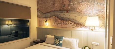 Habitación de hotel con un mapa grande en la pared, una cama con ropa blanca, una mesita de noche con una lámpara y una ventana con vistas.
