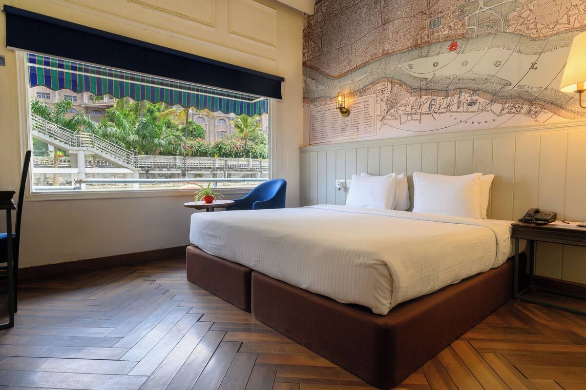 A hotel room with a large bed, a window with a view of palm trees and a building, a map on the wall, and a small table with a chair.
