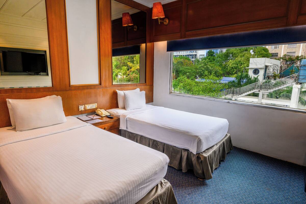 A hotel room with two beds, a TV, a window with a view of greenery and buildings, and a wooden ceiling.