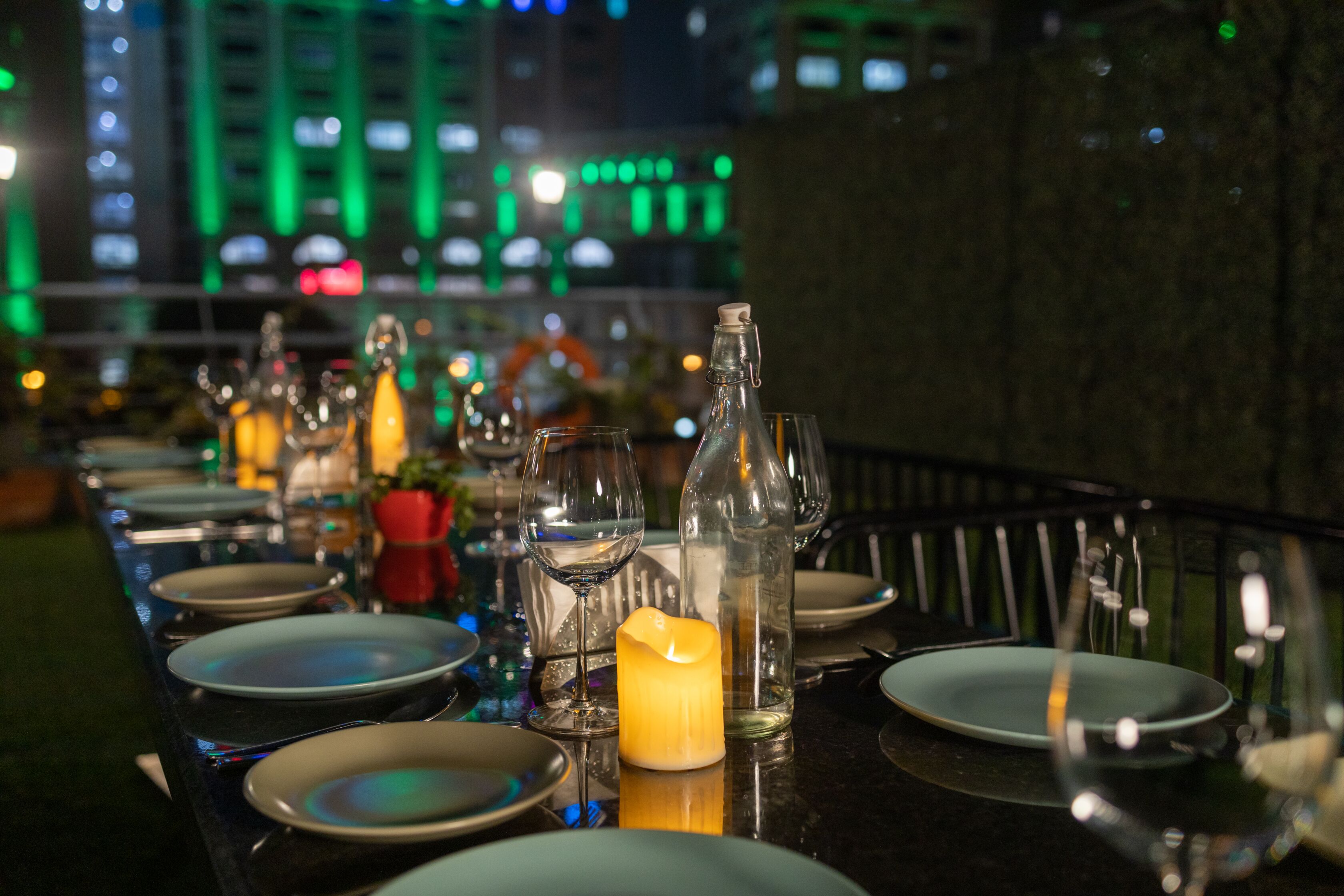 A table set for dining with plates, glasses, and candles, set against a cityscape at night.
