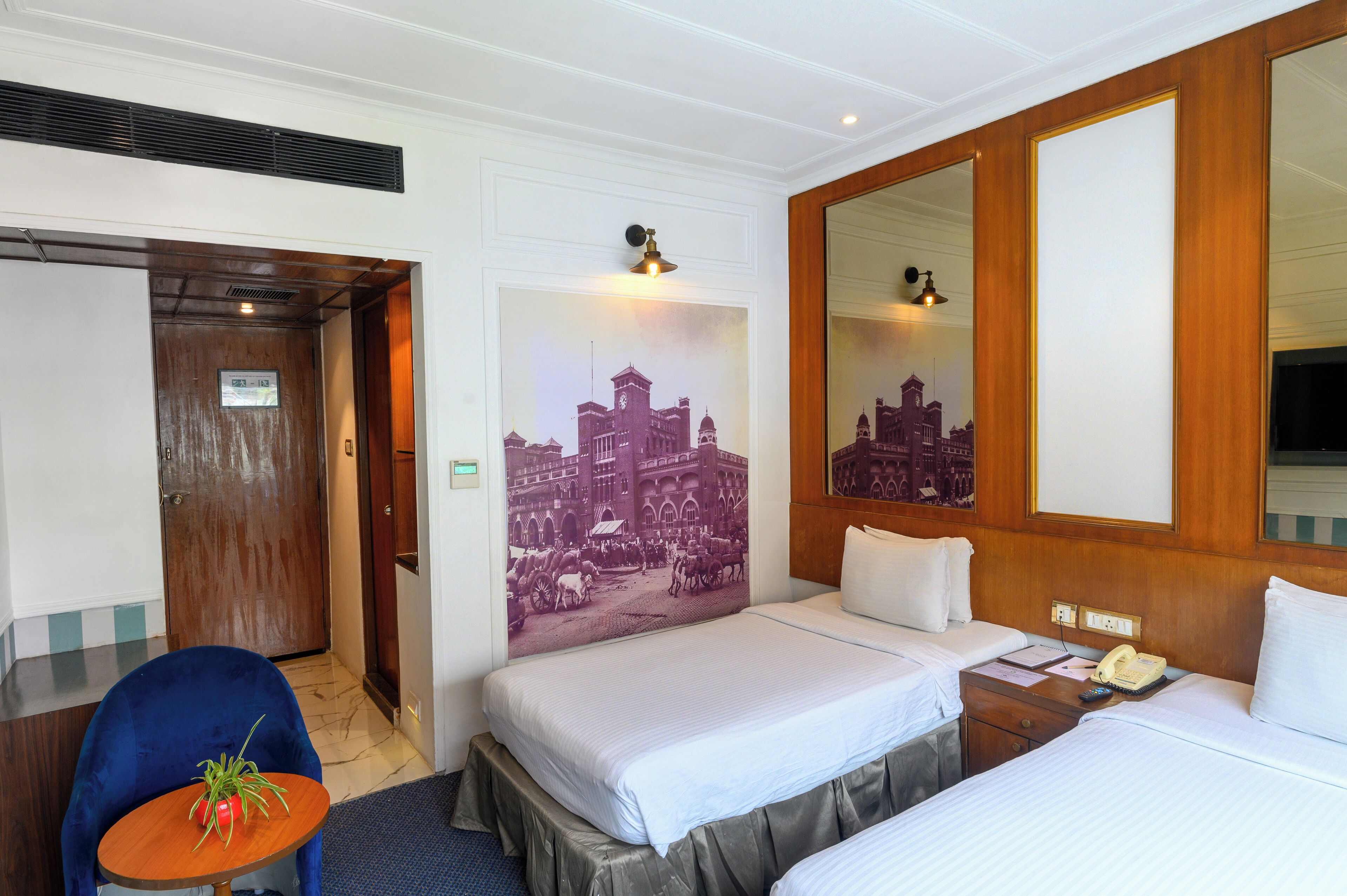 A hotel room with two beds, a wooden headboard, a large mirror, a small table with a plant, and a wall-mounted picture.