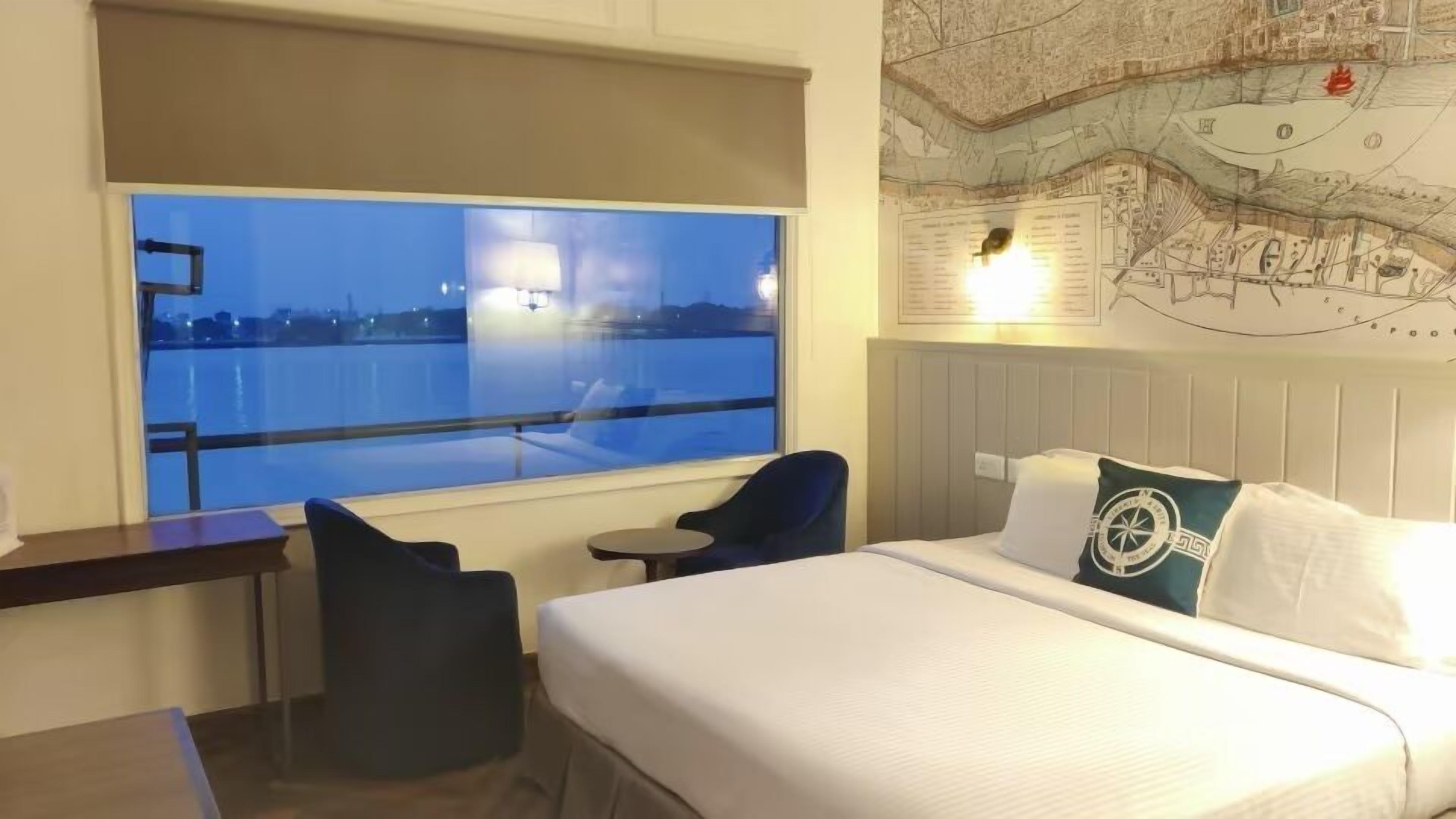 A hotel room with a large window, a bed with white bedding, a map on the wall, and a small table.