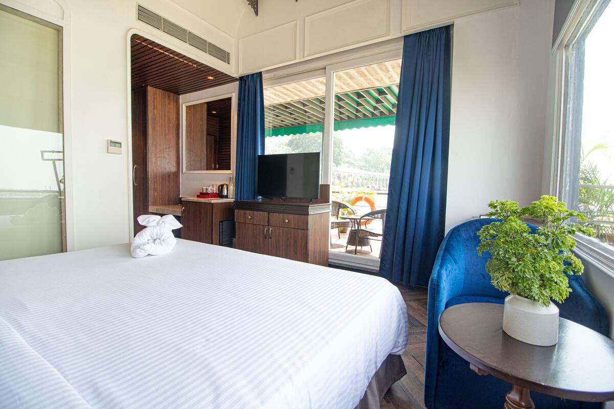 A hotel room with a bed, a desk with a TV, a chair, and a potted plant.