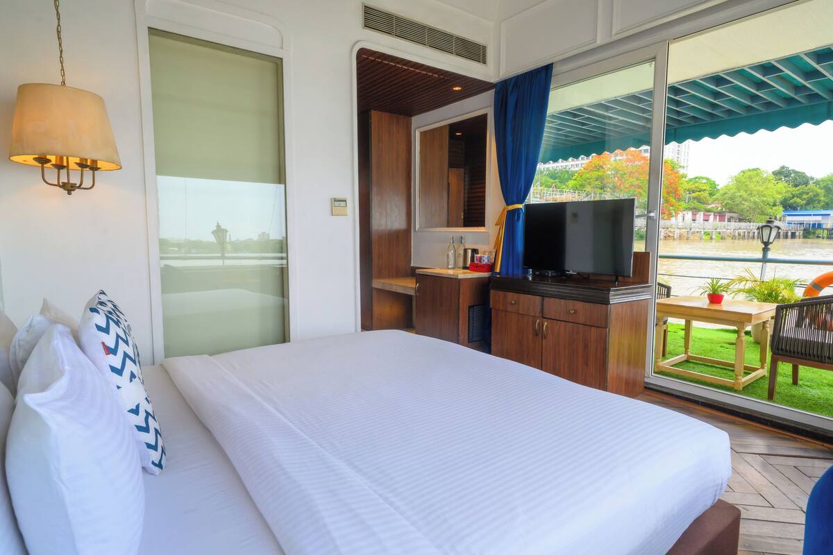A hotel room with a large bed, a television, a desk with a chair, and a view of a river and buildings.