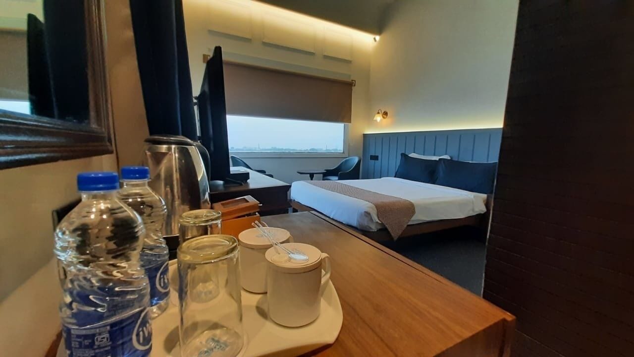 A hotel room with a bed, a desk with a chair, a window, and a table with water bottles and glasses.