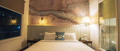 Una habitación de hotel con un mapa grande en la pared, una cama con ropa blanca, una mesita de noche con una lámpara y una ventana con vistas.