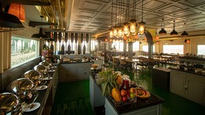 Amplia zona de comedor de hotel con una larga barra de buffet, numerosas mesas y una variedad de alimentos expuestos en una isla central.