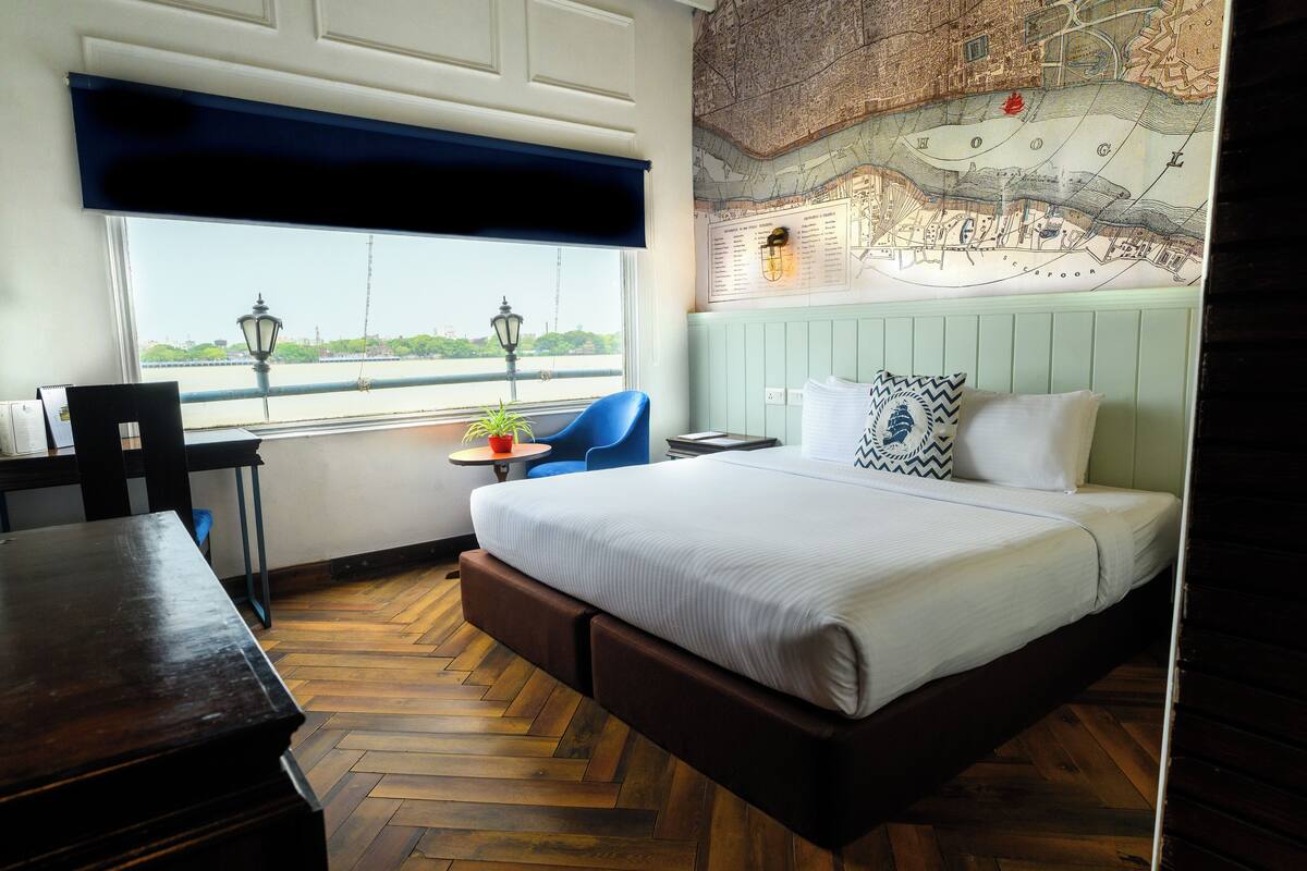 A hotel room with a large bed, a desk, a chair, a small table with a plant, and a view of a river through a window.