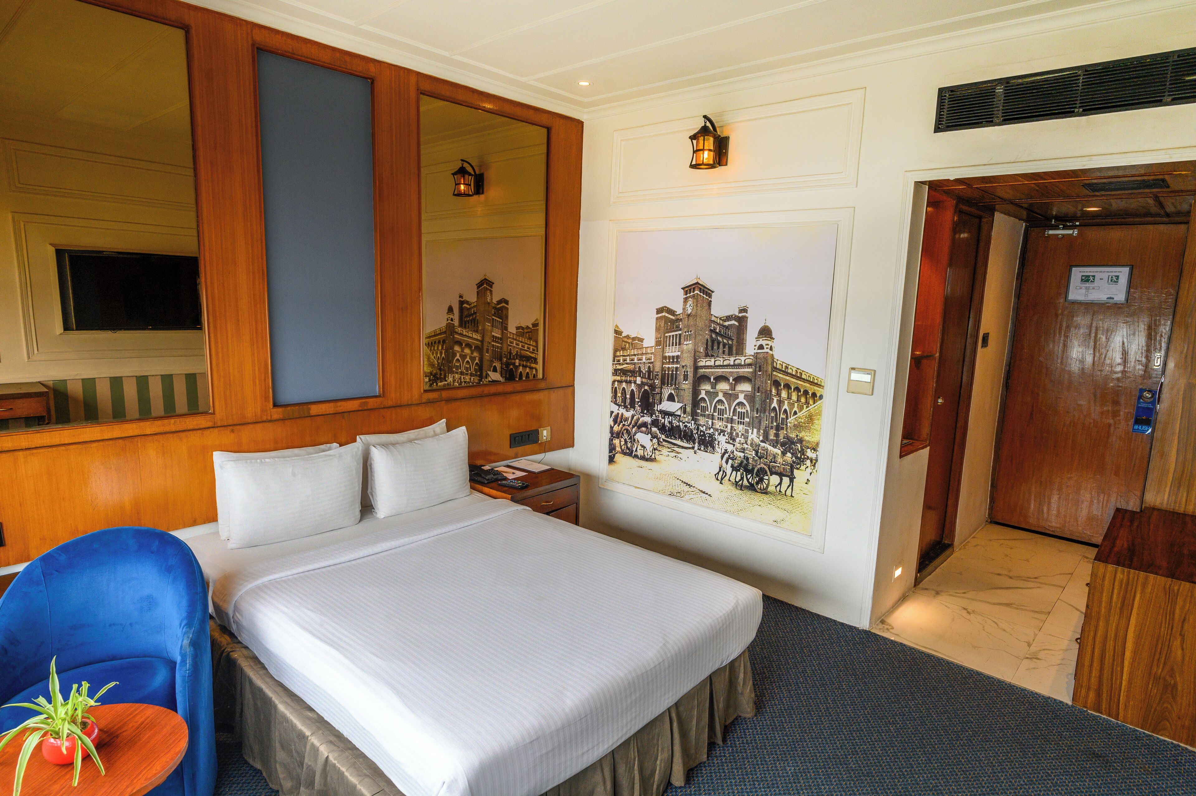 Een hotelkamer met een groot bed, een blauwe stoel, een klein tafeltje met een plant, een spiegel, een wandlamp en een deur naar een lift.