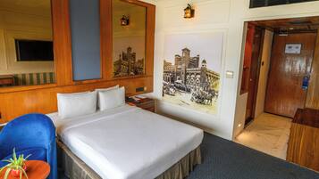 Een hotelkamer met een groot bed, een blauwe stoel, een klein tafeltje met een plant, een spiegel, een wandlamp en een deur naar een lift.