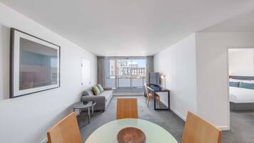 A modern hotel room with a dining area, a sofa, a TV, and a balcony with a city view.