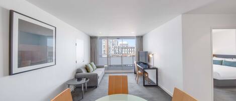 Una habitación de hotel moderna con zona de comedor, sofá, televisión y balcón con vistas a la ciudad.